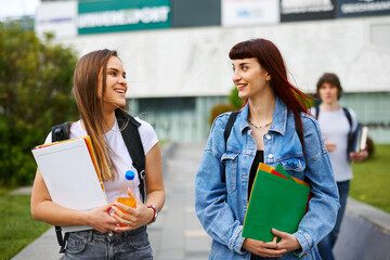 Two happy female students carrying books and backpacks are walking and chatting on a university campus