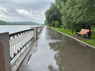 Naklejka premium A park after rain on a cloudy day. A wet red brick path winds through lush green trees and grass. Benches and a gazebo are visible in the background. 