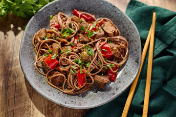 Wok with Soba noodles with turkey, mushrooms and sweet red bell pepper in plate on wooden table in the sun. Asian food, concept of street food. Top view.
