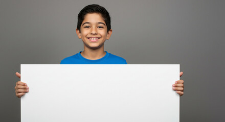 A smiling boy in a blue shirt holding a blank white sign against a solid gray background in a studio shot