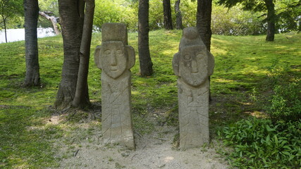 Two stone jangseung figures with distinct facial carvings placed between trees in a grassy area.
