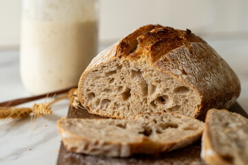 Crispy sourdough bread sits beside a glass jar filled with active starter, accompanied by a rustic cloth and a wooden board in a warm kitchen atmosphere.