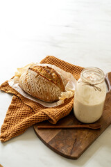 Crispy sourdough bread sits beside a glass jar filled with active starter, accompanied by a rustic cloth and a wooden board in a warm kitchen atmosphere.