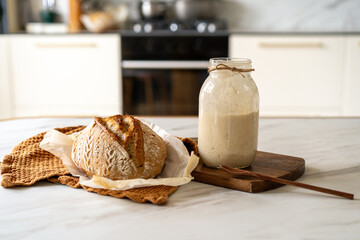 Crispy sourdough bread sits beside a glass jar filled with active starter, accompanied by a rustic cloth and a wooden board in a warm kitchen atmosphere.