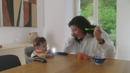 Mother feeding baby at dining table, baby smiling with excitement, joyful interaction between...