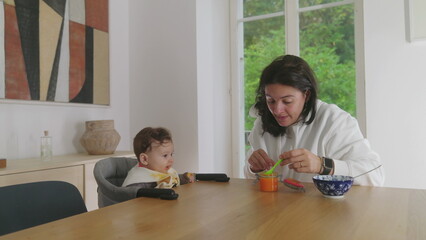 Mother feeding baby at the dining table, baby expressing discomfort while being fed, mother gently...