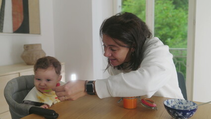 Mother feeding baby at the dining table, baby expressing discomfort while being fed, mother gently...