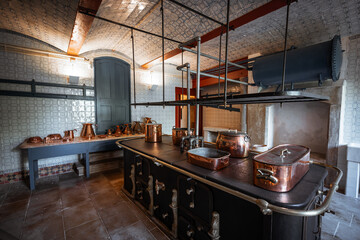 Low-key wide-angle shot of vintage royal kitchen interior with antique copper pots and pans on a black cast iron stove, tiled walls and ceilings, and warm subdued light creating a historical ambiance