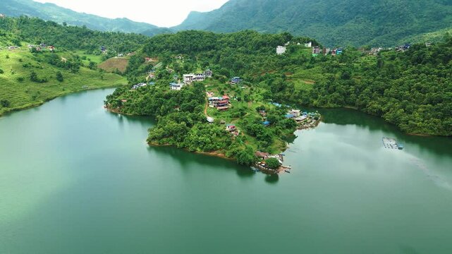 A aerial view of a green piece of land sticking out into a calm lake, with houses, lots of trees, and hills in the background.