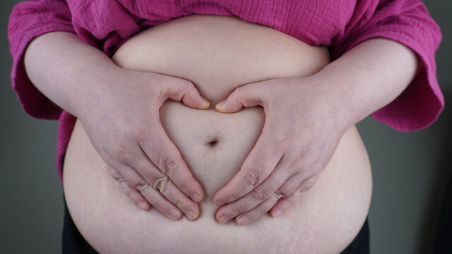 A plump woman shows heart with hands on her fat stomach. A woman in a pink shirt folds her hands on her stomach in the shape of a heart, expressing acceptance of her body and self-love.
