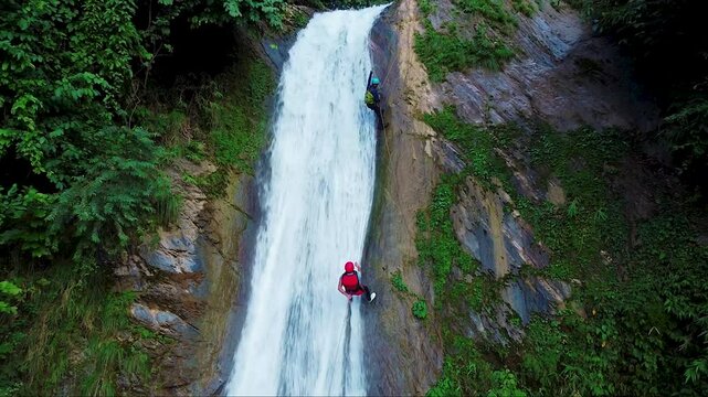 Lush green forest with a cascading waterfall flowing down a rocky cliff. Two adventurers in colorful gear are seen climbing or rappelling along the waterfall.