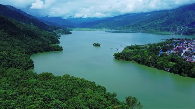 Aerial view of a tranquil lake with a small island, nestled between dense forests and towering mountains under a cloudy sky. 