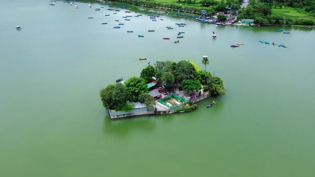 Aerial shot of a small, lush island in a lake, surrounded by boats and paddle boats, with dense greenery.