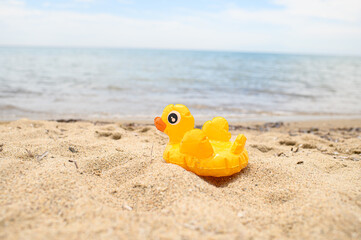 Obraz premium Yellow inflatable duck toy lying on sandy beach near the sea. Bright summer concept, vacation vibes, seaside fun, minimal beach background with copy space.