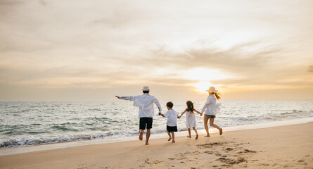 A happy Asian family seen from behind enjoys sunny day at beach. Parents and daughters bond running jumping and pretending to fly with arms outstretched. carefree family moment by sea.