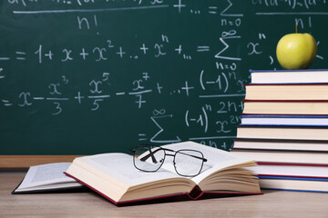 Books, glasses and apple on wooden table near blackboard with formulas, closeup
