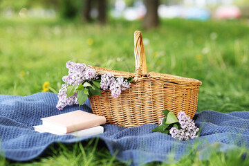 Stacked books, blanket and wicker basket with lilac flowers on green grass outdoors, closeup