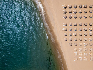 Aerial top-down view of a sandy beach with straw parasols and sunbeds aligned in rows, waves gently washing ashore. Summer vacation concept