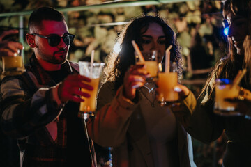 Friends gathered together at a lively celebration, clinking glasses of cocktails under party lights.