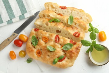 Cut delicious focaccia with tomatoes, basil, oil and knife on light table, flat lay