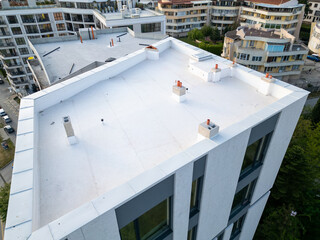 Aerial view of modern residential buildings under construction, featuring flat white rooftops, scaffolding, and unfinished facades. Surrounding neighborhood with completed homes in background