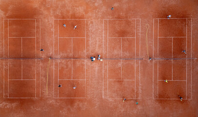 Aerial top-down view of multiple red clay tennis courts with players practicing. Symmetrical sports layout with lines, nets, and visible training activity on a sunny day