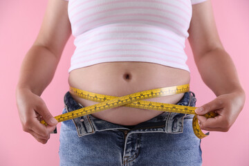 Plakat Overweight woman measuring her belly with tape on pink background, closeup