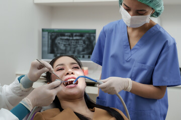 Asian female dentist and assistant using dental suction and mirror while treating adult female patient during oral checkup procedure in dental clinic with digital x-ray on monitor.
