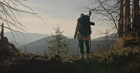 Rear view of girl in hiking clothes with big backpack stepping on hill and looking at forests and mountains during her trip at sunrise