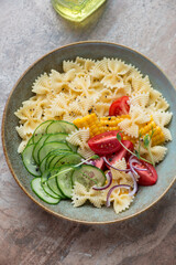 Rustic plate with farfalle, cucumber, tomatoes and sweet corn pasta salad, vertical shot on a brown granite background, high angle view