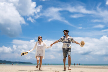 Couple walks hand in hand along sandy beach under bright clouds on sunny day