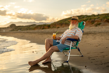 Relaxed senior man with white beard and straw hat sitting in a beach chair by the sea, holding orange juice, enjoying peaceful sunset moments and retirement lifestyle on a calm summer evening.