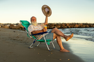 Cheerful elderly man relaxing on the beach, sitting in a chair with a glass of orange juice, smiling and waving his straw hat, enjoying retirement, freedom and happiness during golden sunset.