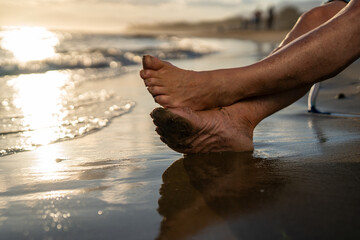 Close-up of bare senior feet resting on wet sandy beach at sunset, one covered in sand, waves gently reaching shoreline, peaceful moment of barefoot relaxation by the sea...
