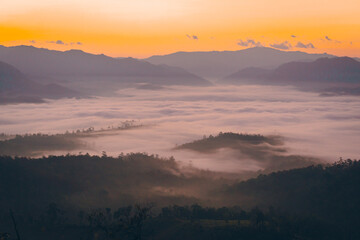 Obraz premium High angle view of the mountain with fog in the morning in Thailand. The sun rises above clouds in warm tones. The idea for a nature background with copy space. The fog is very thick and fluffy.
