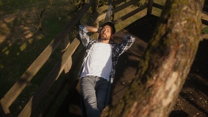 View from above of handsome Caucasian male resting after hard working day at farm. Laying in hammock under tall tree. Spending free time in fresh air on deck. Evening coming in village.