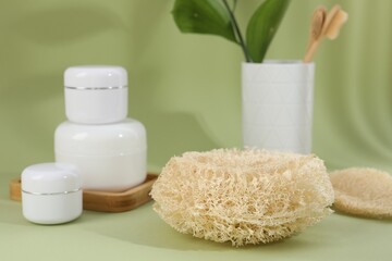 Loofah sponge and jars of cosmetic product on olive background, closeup