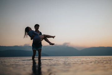 Fototapeta premium A man holds a woman in his arms while standing in the calm waters of a lake during sunset, evoking a scene of romance and tranquility amidst a tranquil natural setting.