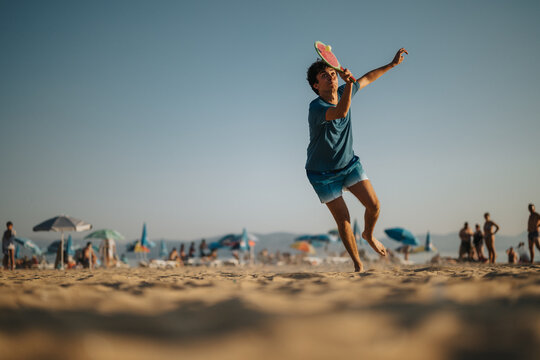 A young man energetically plays beach ball on the sandy shore under a sunny sky. Umbrellas and people enjoying their leisure surround the scene, creating a summer recreational feeling.