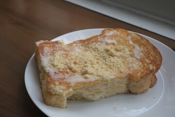 Close-up of a slice of golden brown toast topped with sweet condensed milk and powdered crumbs on a white plate, served on a wooden table
