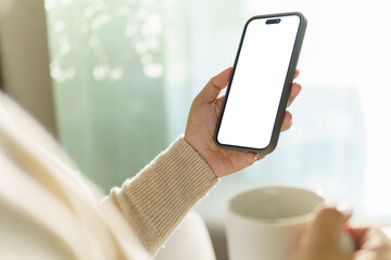 Hand holding a smartphone with a blank screen while enjoying a cup of coffee, for app mockups or...