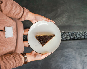 Hands holding a white plate with a tempting piece of layered cake, with chocolate crumbs and cream filling. A delicious dessert