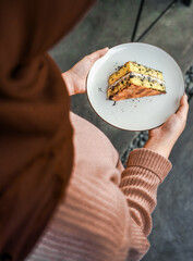 Hands holding a white plate with a tempting piece of layered cake, with chocolate crumbs and cream filling. A delicious dessert
