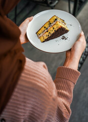 Hands holding a white plate with a tempting piece of layered cake, with chocolate crumbs and cream filling. A delicious dessert