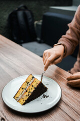 A person cuts into a delicious slice of layered cake with chocolate crumbs and creamy filling, served on a white plate on a wooden table