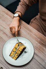 A person cuts into a delicious slice of layered cake with chocolate crumbs and creamy filling, served on a white plate on a wooden table
