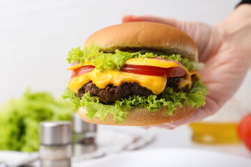 Woman holding tasty cheeseburger at white table, closeup