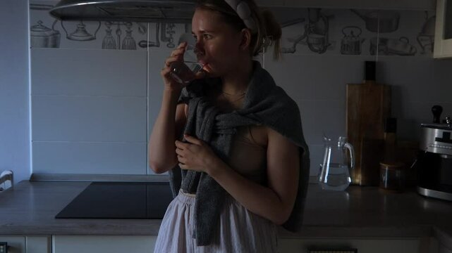 A woman stands in a warmly lit kitchen, sipping from a glass as she wears a sweater over a light outfit. The atmosphere is relaxed and intimate, perfect for winding down.