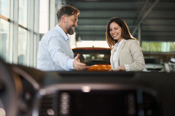 Happy car salesman showing a car to his female customer in a showroom. The view is through...