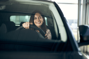 Happy woman enjoying  inside of a car in a showroom. The view is through windshield.